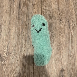 May include: A light green, soft, crocheted plush toy shaped like a pickle. The toy features a textured, yarn-based surface with two small, round, black plastic eyes and a simple, stitched black V-shaped mouth centred on the front side.