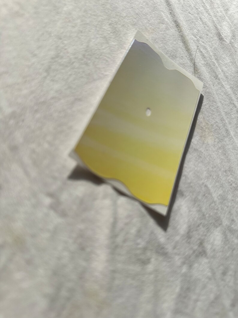 May include: A small, rectangular, silver-colored sticker with a yellow gradient and a small hole in the center.