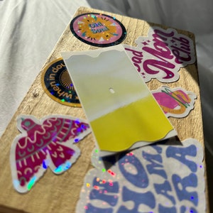 May include: A wooden box with various colorful stickers, including a sticker with the text "Old Soul", a sticker with the text "Bad Mom Club", a sticker with a bird design, and a sticker with the text "I'm Home Mama" in blue and purple.