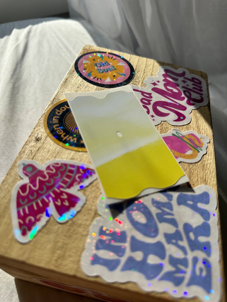 May include: A wooden box with various colorful stickers, including a sticker with the text "Old Soul", a sticker with the text "Bad Mom Club", a sticker with a bird design, and a sticker with the text "I'm Home Mama" in blue and purple.