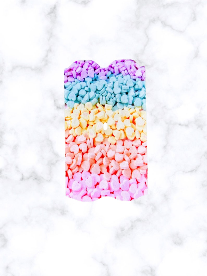 May include: A rainbow-colored assortment of heart-shaped sprinkles in a rectangular container. The sprinkles are arranged in rows of purple, blue, yellow, orange, and pink.