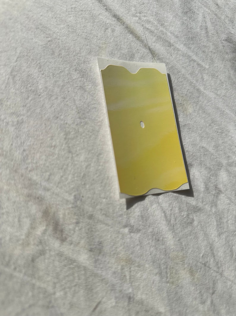May include: A gold-colored, rectangular sticker with a wavy edge and a small hole in the center.
