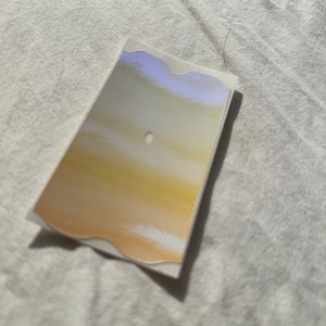 May include: A white sticker with a wavy edge and a gold and white gradient design. The sticker has a small hole in the center.