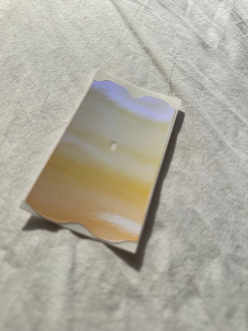 May include: A white sticker with a wavy edge and a gold and white gradient design. The sticker has a small hole in the center.