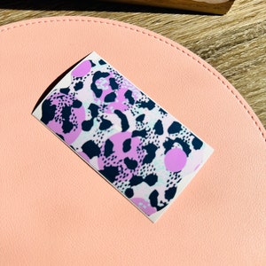 May include: A sticker featuring an abstract animal print design in pink, purple and black. The print has a leopard-like pattern with large, round spots.