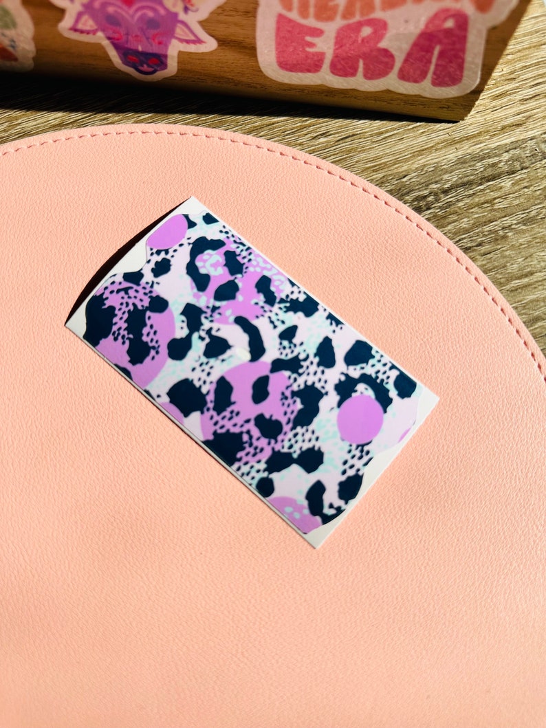 May include: A sticker featuring an abstract animal print design in pink, purple and black. The print has a leopard-like pattern with large, round spots.