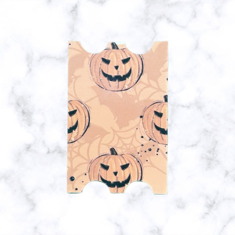May include: A peach-colored card with a scalloped edge features a repeating pattern of orange jack-o'-lantern faces with black eyes and mouths. The background has a subtle spiderweb pattern.