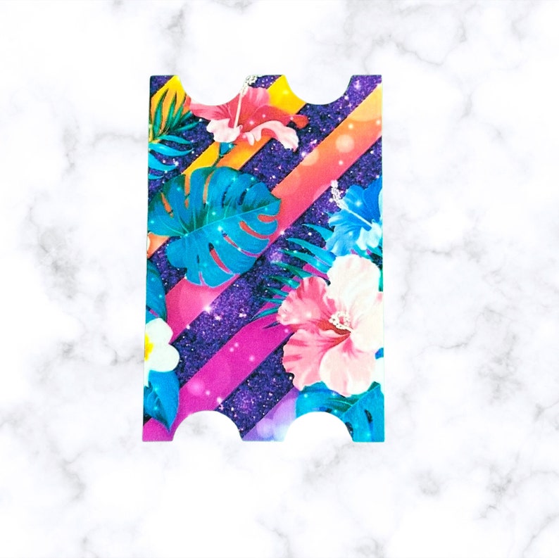 Pu&ograve; includere: Un biglietto colorato a tema tropicale. Il biglietto presenta fiori di ibisco rosa, blu e gialli, foglie verdi e uno sfondo glitterato viola e rosa.