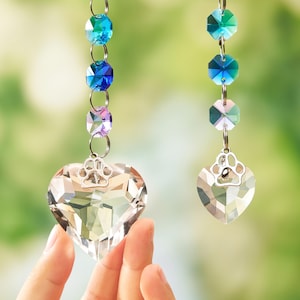 May include: Two hanging ornaments featuring clear faceted crystal hearts topped with silver-toned metal paw print charms. Each heart is suspended from a vertical chain of multi-colored faceted crystal beads in shades of blue, teal, and purple.