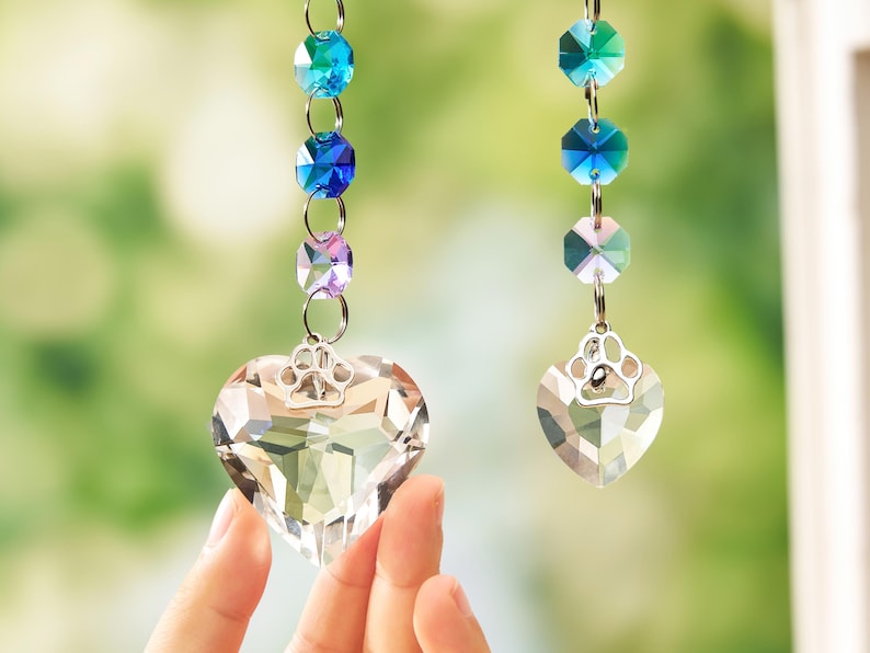 May include: Two hanging ornaments featuring clear faceted crystal hearts topped with silver-toned metal paw print charms. Each heart is suspended from a vertical chain of multi-colored faceted crystal beads in shades of blue, teal, and purple.