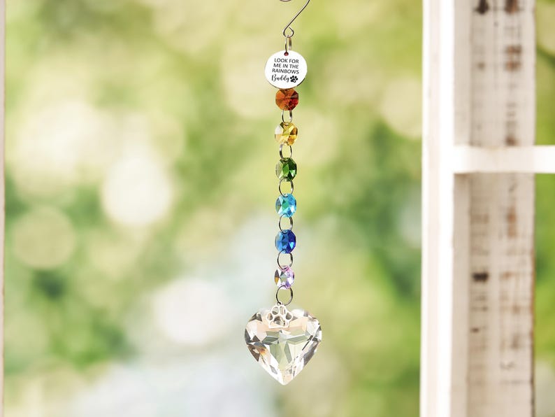 May include: A hanging suncatcher ornament featuring a circular silver charm with the text LOOK FOR ME IN THE RAINBOWS Buddy and a paw print. Below the charm are six faceted crystal beads in rainbow colors, ending in a clear heart-shaped crystal.