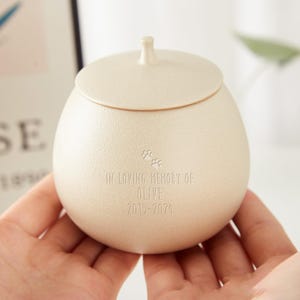 Puede incluir: Una peque&ntilde;a urna de cer&aacute;mica de color crema con tapa. Tiene un dise&ntilde;o de huella de pata y la inscripci&oacute;n "In loving memory of Olive 2015-2021".