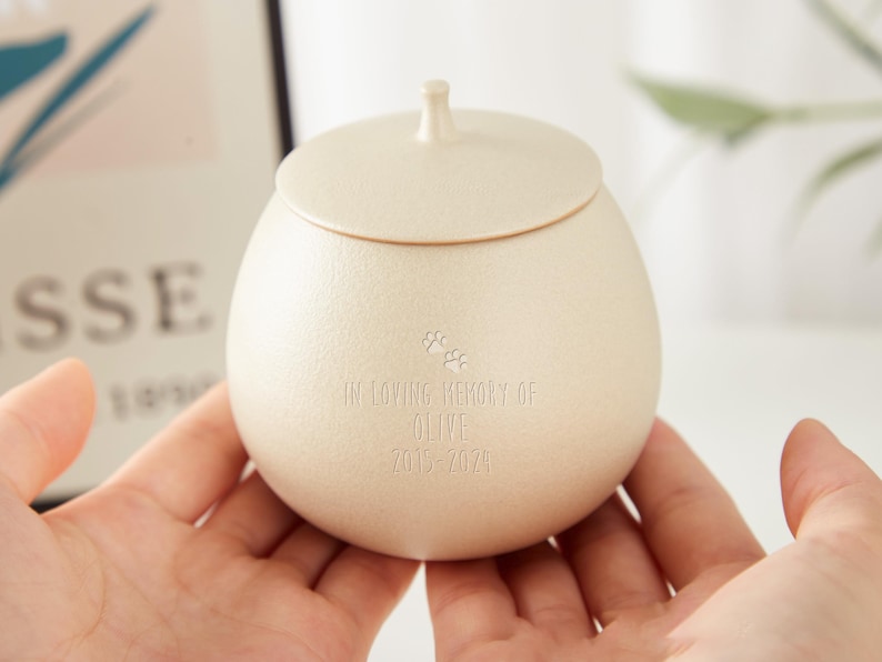 Puede incluir: Una peque&ntilde;a urna de cer&aacute;mica de color crema con tapa. Tiene un dise&ntilde;o de huella de pata y la inscripci&oacute;n "In loving memory of Olive 2015-2021".