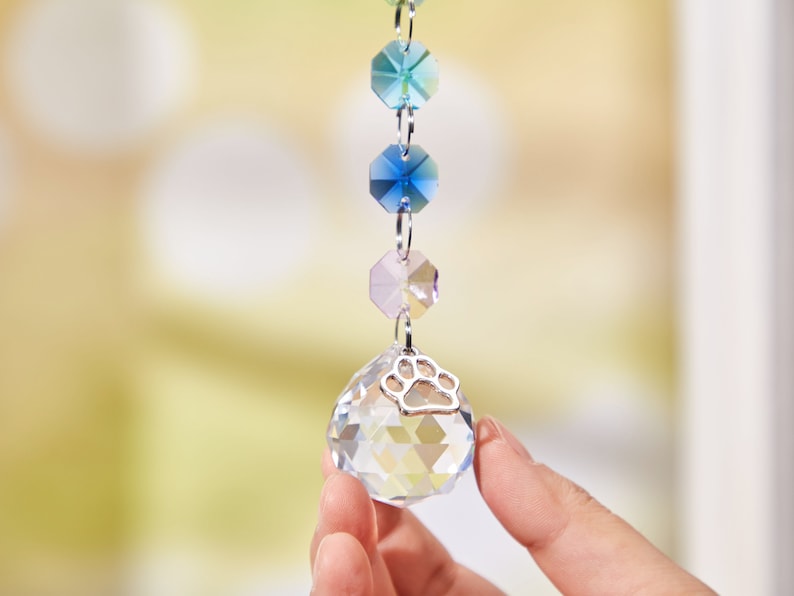 May include: A crystal suncatcher with a silver paw print charm. The suncatcher features a clear crystal ball with faceted sides and a chain of iridescent blue, green, and pink crystals.