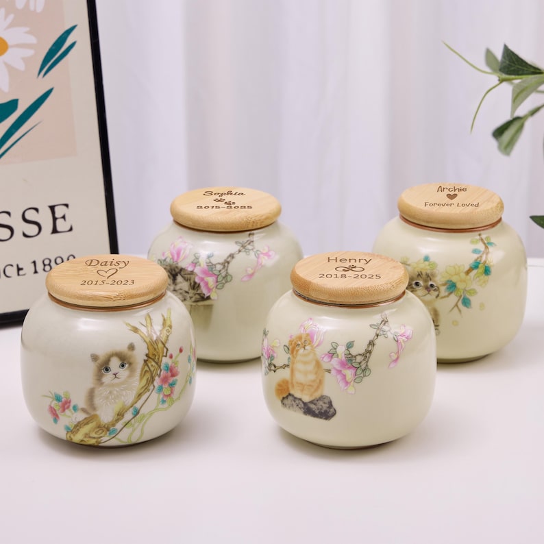 May include: Four ceramic pet urns with wooden lids. Each urn features a painted cat illustration and floral accents in pink and green. The lids are engraved with names and dates, including "Daisy," "Sophia," "Henry," and "Archie."