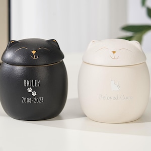 Peut inclure: Deux urnes en forme de chat, une noire et une crème. L'urne noire porte le nom "Bailey", des empreintes de pattes et les années "2008-2023". L'urne crème dit "Beloved Coco". Les deux ont des accents dorés et des visages souriants.