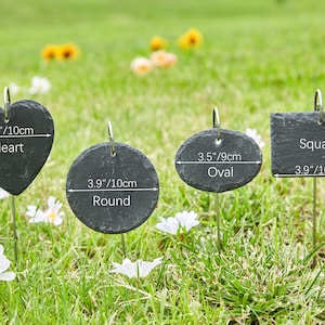 May include: Four black slate plant markers with different shapes: heart, round, oval, and square. Each marker has a metal stake and includes measurements in inches and centimeters.
