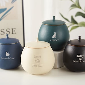 Puede incluir: Cuatro urnas de cer&aacute;mica para mascotas con tapas en diferentes colores: azul, crema, verde y negro. Cada urna tiene una inscripci&oacute;n personalizada con el nombre de la mascota y un peque&ntilde;o gr&aacute;fico de una huella de pata o un animal. Las inscripciones dicen: "Beloved Coco", "Wally 2005-2023", "Copper" y "Kitty Forever Loved".