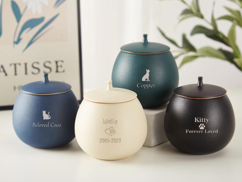Puede incluir: Cuatro urnas de cer&aacute;mica para mascotas con tapas en diferentes colores: azul, crema, verde y negro. Cada urna tiene una inscripci&oacute;n personalizada con el nombre de la mascota y un peque&ntilde;o gr&aacute;fico de una huella de pata o un animal. Las inscripciones dicen: "Beloved Coco", "Wally 2005-2023", "Copper" y "Kitty Forever Loved".