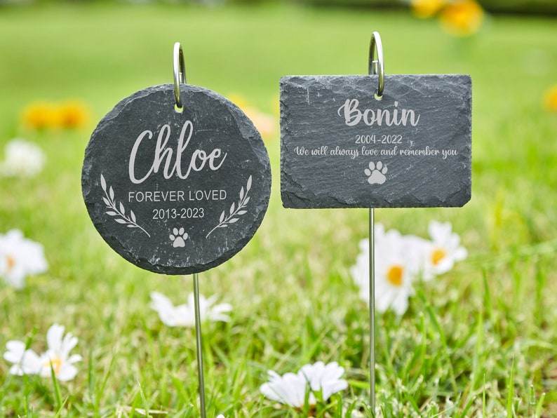 May include: Two black slate pet memorial markers with engraved text. The first marker is round and says "Chloe Forever Loved 2013-2023" with a paw print. The second marker is rectangular and says "Bonin 2004-2022 We will always love and remember you" with a paw print.