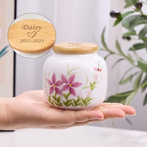 May include: A white ceramic urn with a wooden lid, hand-painted with pink flowers and green leaves. The lid is engraved with the name "Daisy", a heart symbol, and the years 2015-2023. A dragonfly is also painted on the urn.