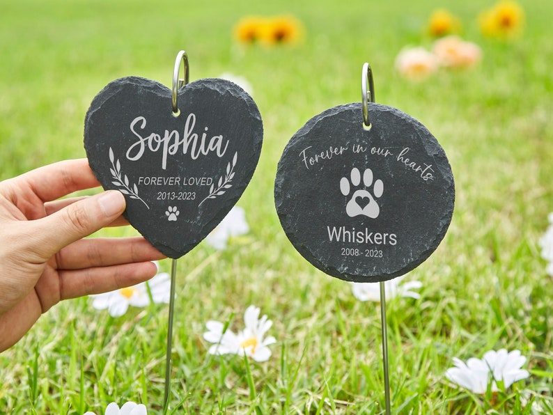 May include: Two slate garden markers, one heart-shaped and one round. The heart-shaped marker reads "Sophia Forever Loved 2013-2023" with a paw print. The round marker reads "Forever in our hearts" with a paw print and "Whiskers 2008-2023".