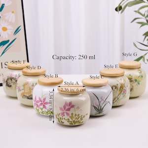 May include: A collection of ceramic jars with wooden lids, each adorned with floral or cat-themed designs. The jars are approximately 3.34 inches (8.5 cm) tall and 2.36 inches (6 cm) in diameter, with a capacity of 250 ml.