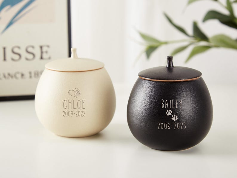 Puede incluir: Dos urnas para mascotas de cer&aacute;mica con tapas. La urna de la izquierda es de color crema con el nombre Chloe y las fechas 2009-2023. La urna de la derecha es negra con el nombre Bailey y las fechas 2008-2023.