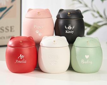 May include: Five ceramic cat-shaped urns in red, pink, white, black, and green. Each urn features a smiling cat face on the lid and a name or design on the front. These urns are designed for pet memorial purposes.