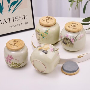 May include: Four ceramic pet urns with wooden lids. The urns are off-white with floral and cat illustrations. The lids are engraved with names and the phrase "Forever Loved". One lid is off and shows a gray interior.