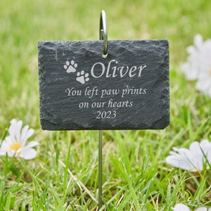 May include: A black slate garden marker with the text "Oliver" and "You left paw prints on our hearts 2023". The marker has two paw prints and is attached to a metal stake.