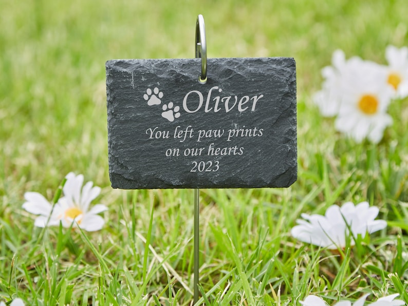 May include: A black slate garden marker with the text "Oliver" and "You left paw prints on our hearts 2023". The marker has two paw prints and is attached to a metal stake.