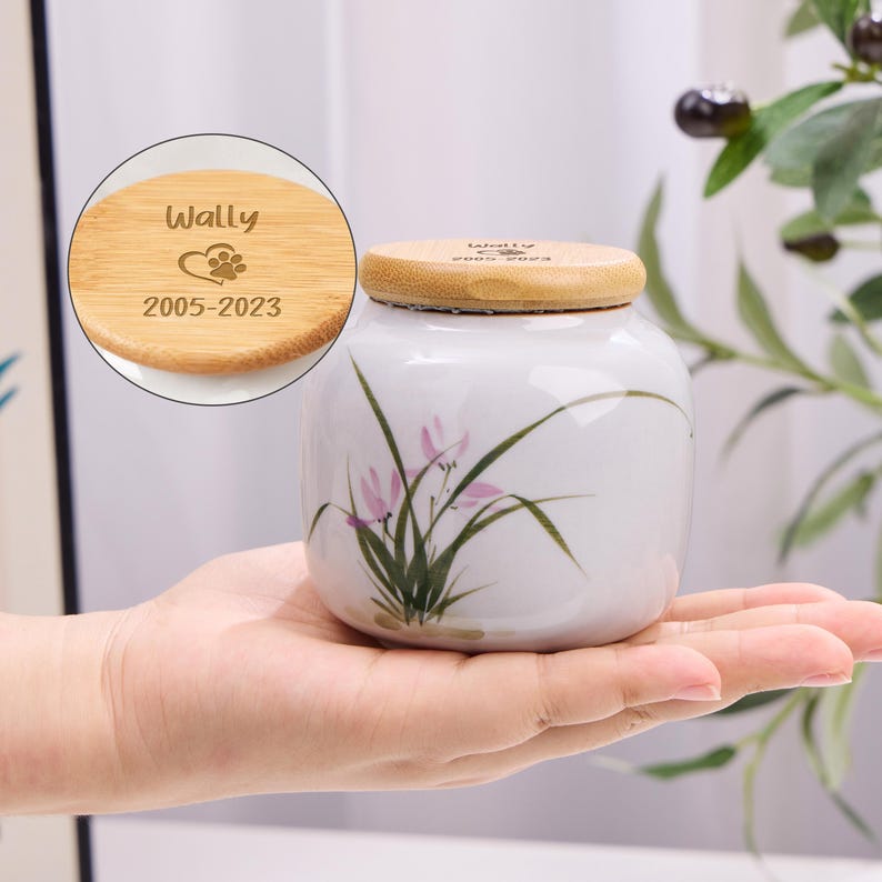 May include: A white ceramic pet urn with a bamboo lid. The urn is decorated with a floral design and held in a hand. The lid is engraved with the name "Wally", a heart and paw print, and the years "2005-2023".