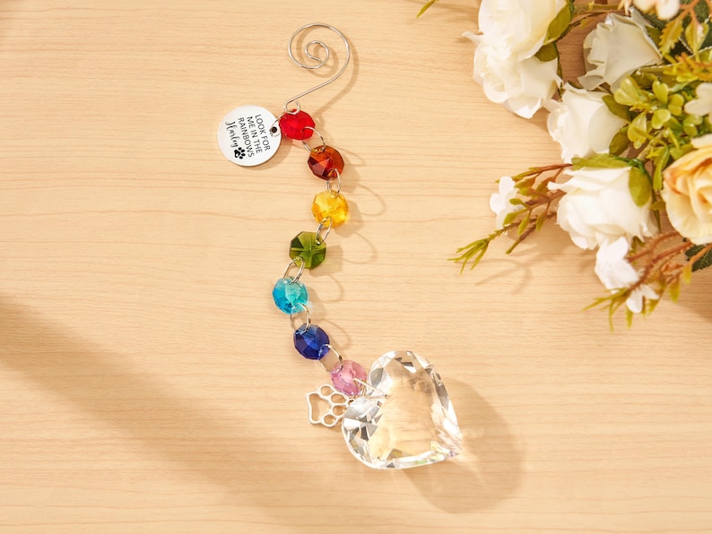 May include: A hanging ornament featuring a silver spiral hook, a round metal tag engraved with "LOOK FOR ME IN THE RAINBOWS" and a paw print, a vertical strand of multi-colored faceted crystal beads, a silver paw print charm, and a clear crystal heart.