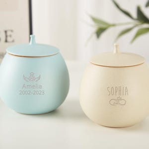 Puede incluir: Dos urnas de cer&aacute;mica con tapas, una azul claro y otra crema. La urna azul tiene el nombre Amelia y las fechas 2002-2023 grabadas. La urna crema tiene el nombre Sophia grabado.