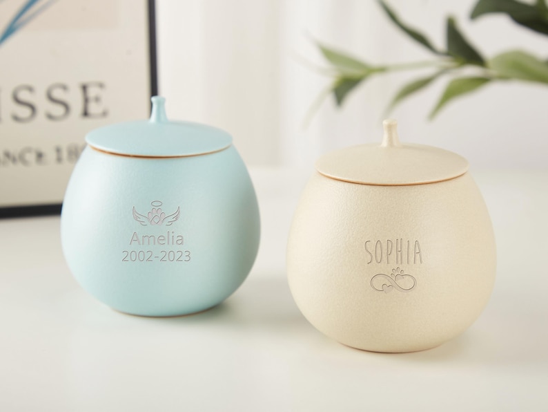 Puede incluir: Dos urnas de cer&aacute;mica con tapas, una azul claro y otra crema. La urna azul tiene el nombre Amelia y las fechas 2002-2023 grabadas. La urna crema tiene el nombre Sophia grabado.