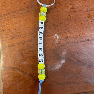 May include: A yellow and blue beaded keychain with the word "FEARLESS" spelled out in white letter beads.