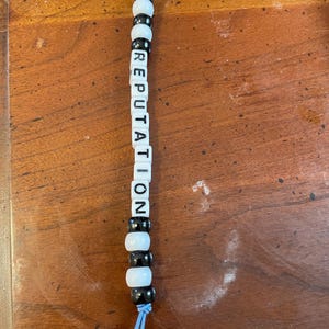 May include: A black and white beaded keychain with the word "REPUTATION" spelled out in black letters on white beads. The keychain has a silver key ring and a blue string.