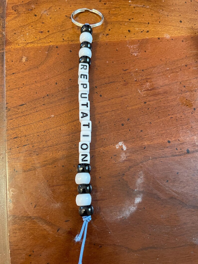 May include: A black and white beaded keychain with the word "REPUTATION" spelled out in black letters on white beads. The keychain has a silver key ring and a blue string.