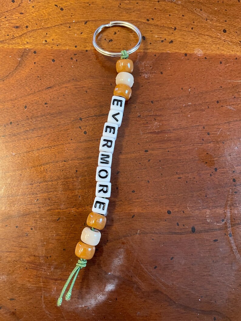 May include: A white and brown beaded keychain with the word "EVERMORE" spelled out in black letters.