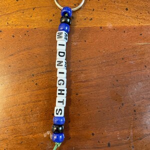 May include: A blue and black beaded keychain with the word "MIDNIGHTS" spelled out in white letter beads.