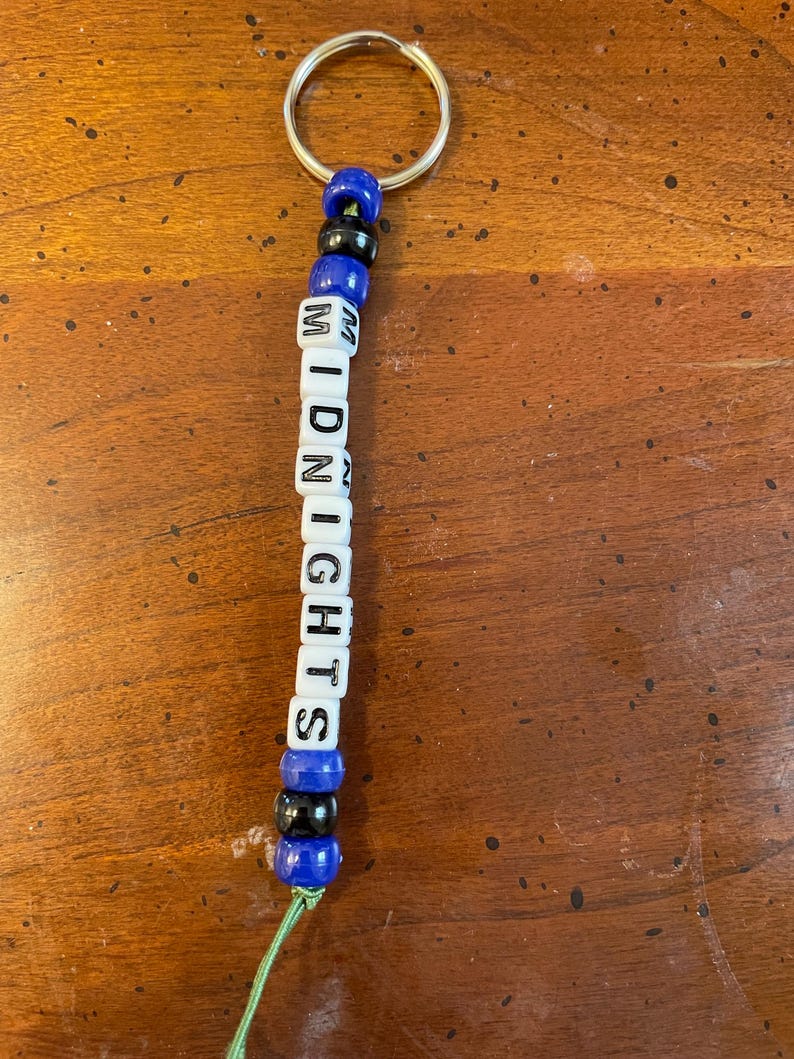 May include: A blue and black beaded keychain with the word "MIDNIGHTS" spelled out in white letter beads.