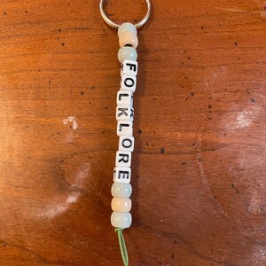 May include: A keychain with a silver ring and a string of beads that spell out "FOLKLORE". The beads are white with black lettering and alternate between light blue and peach.