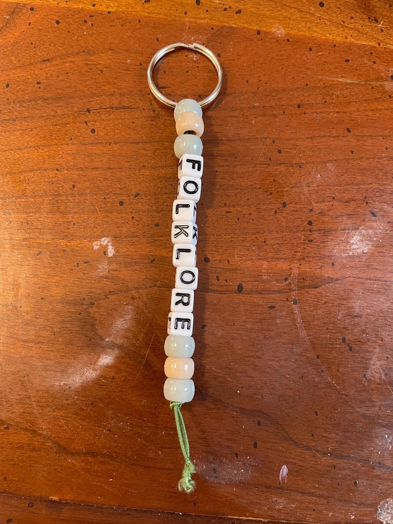 May include: A keychain with a silver ring and a string of beads that spell out "FOLKLORE". The beads are white with black lettering and alternate between light blue and peach.