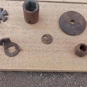 Op de afbeelding: Een verzameling van zes verweerde, roestige metalen onderdelen, waaronder een stervormige ring, een cilindrische dopsleutel, een grote platte ring, een kleine gebogen beugel met een vierkant gat, een kleine ring en een zeskantmoer.