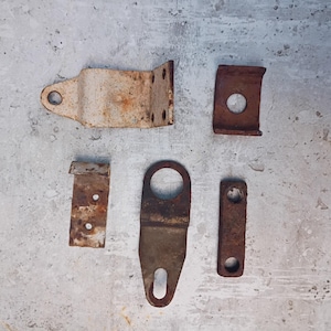 May include: An assortment of five rusty metal brackets and fasteners. The pieces vary in shape, including right angles, curved pieces, and flat bars with holes. The metal has a weathered, aged appearance with visible rust.