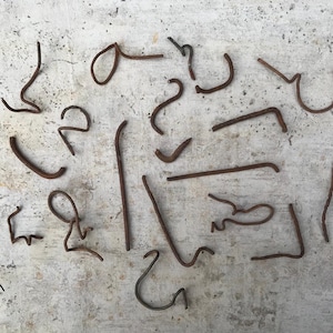 Könnte beinhalten: Eine Ansammlung von rostigen Metallteilen, darunter gebogene Stäbe, Haken und Draht, liegt auf einer Betonoberfläche. Die Formen variieren von einfachen Kurven bis zu komplexeren Formen, alle mit einem verwitterten Aussehen.