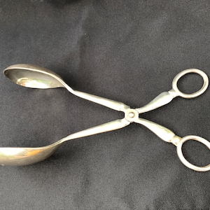 May include: Silver salad tongs with two spoon-shaped ends. The tongs are open and ready to serve.