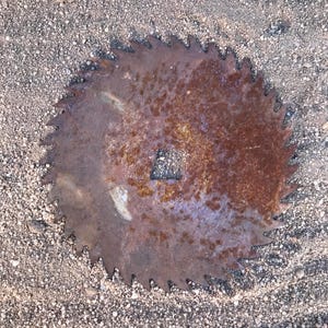 Peut inclure: Une lame de scie circulaire en métal dotée d'un bord dentelé et d'un trou central de forme carrée. La surface de la lame présente une oxydation importante, caractérisée par une patine de rouille brune et orange sur tout le disque métallique.