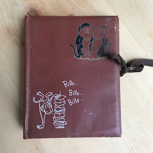 May include: Brown notebook with a ribbon tie closure. The cover features a cartoon illustration of two monkeys, one covering its eyes and the other its ears. Below, a monkey is shown reaching for a stack of bills, with the word "Bills" repeated.